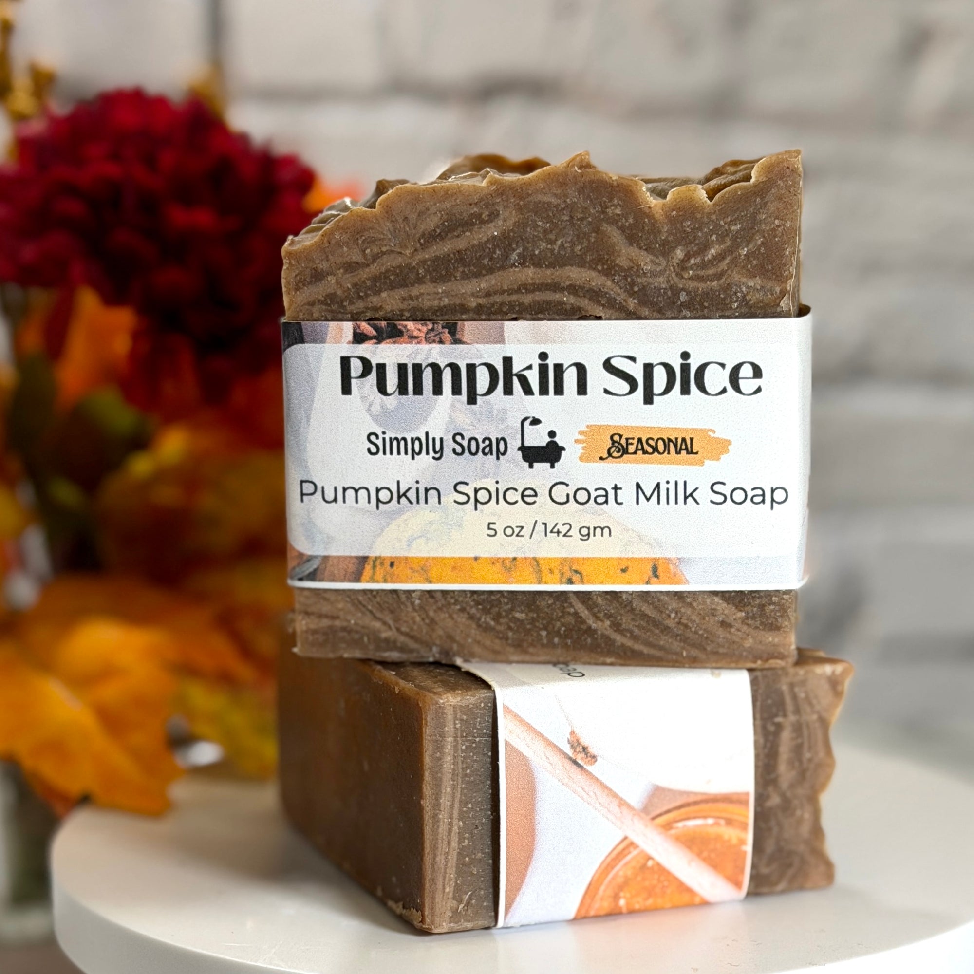 Two stacked bars of Simply Soap Pumpkin Spice Goat Milk Soap with a white paper label, displayed on a white round pedestal with autumn leaves and a white brick wall in the background.