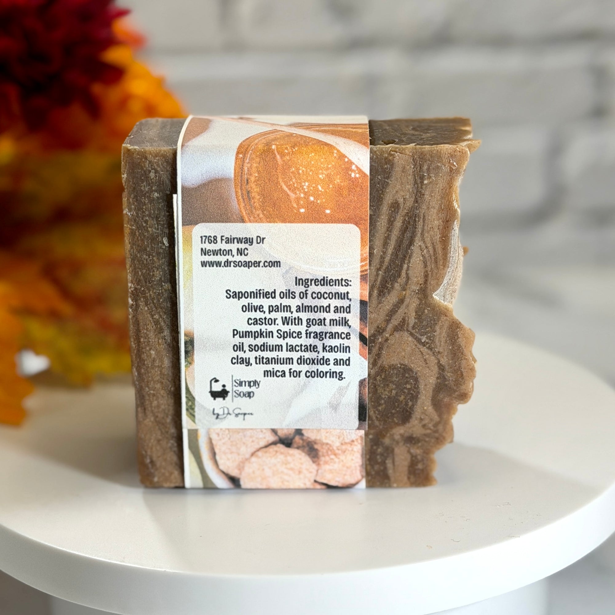 A handmade bar of soap with a brown and beige marbled pattern, wrapped in a paper band label, displayed on a white round pedestal with a blurred background.