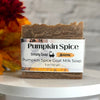 A bar of Simply Soap Pumpkin Spice Goat Milk Soap sits on a white round table. The soap has a rustic, marbled brown texture and is wrapped in a white paper label with black text and orange accents.