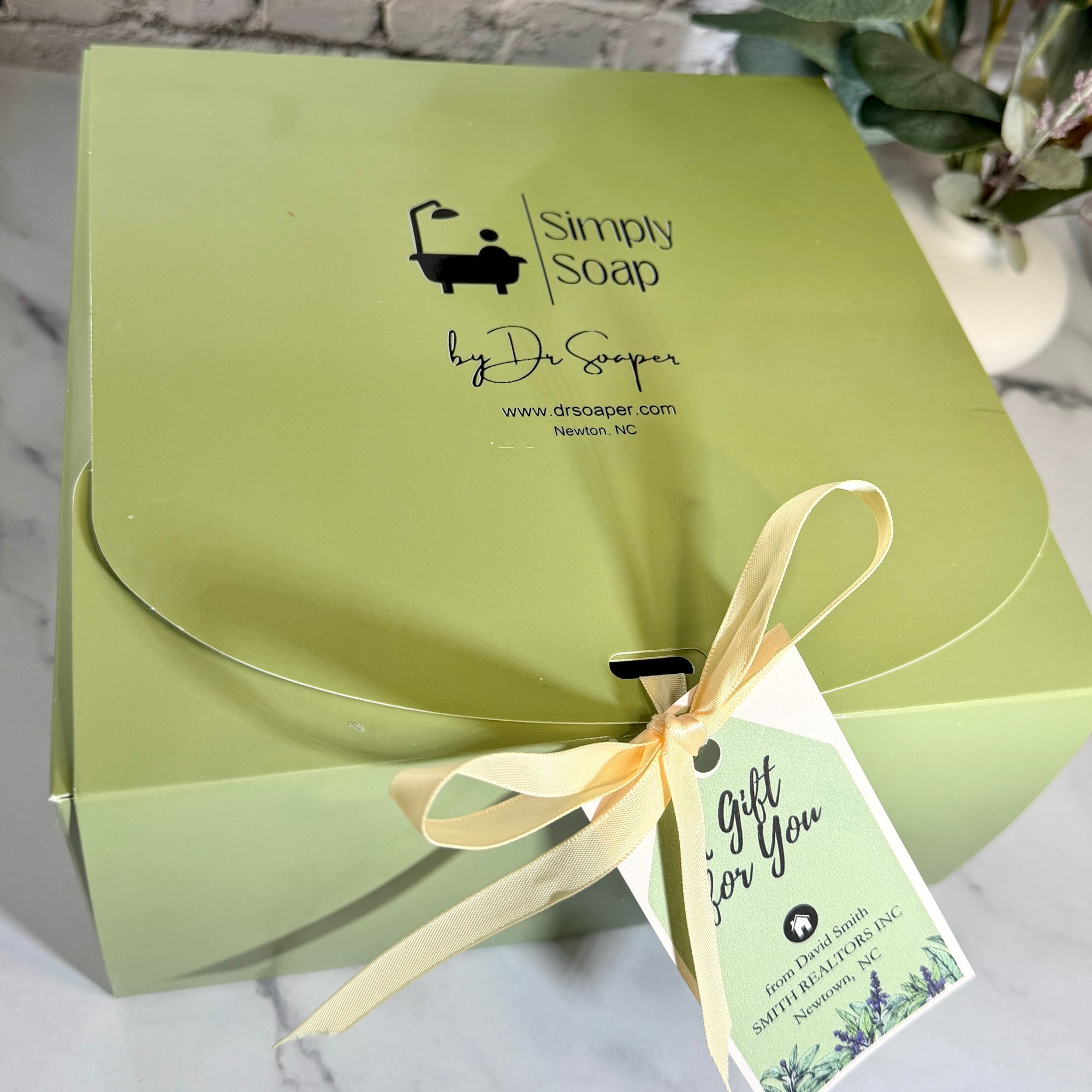 A light green gift box for Simply Soap by Dr. Soaper, tied with a pale yellow ribbon and a 'Gift for You' Realtor tag, set on a marble surface.