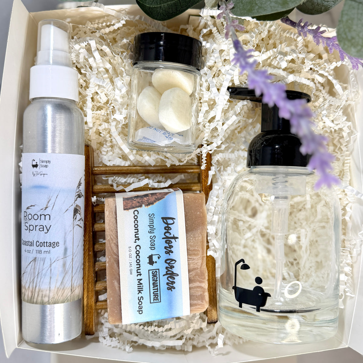 A curated Realtors closing or housewarming gift  arranged in a white box with shredded paper filler. The set includes a silver room spray bottle labeled 'Bloom Cottage', a small jar of white soap tabs, a bar of soap labeled 'Bathhouse Essentials', and a large clear pump bottle.