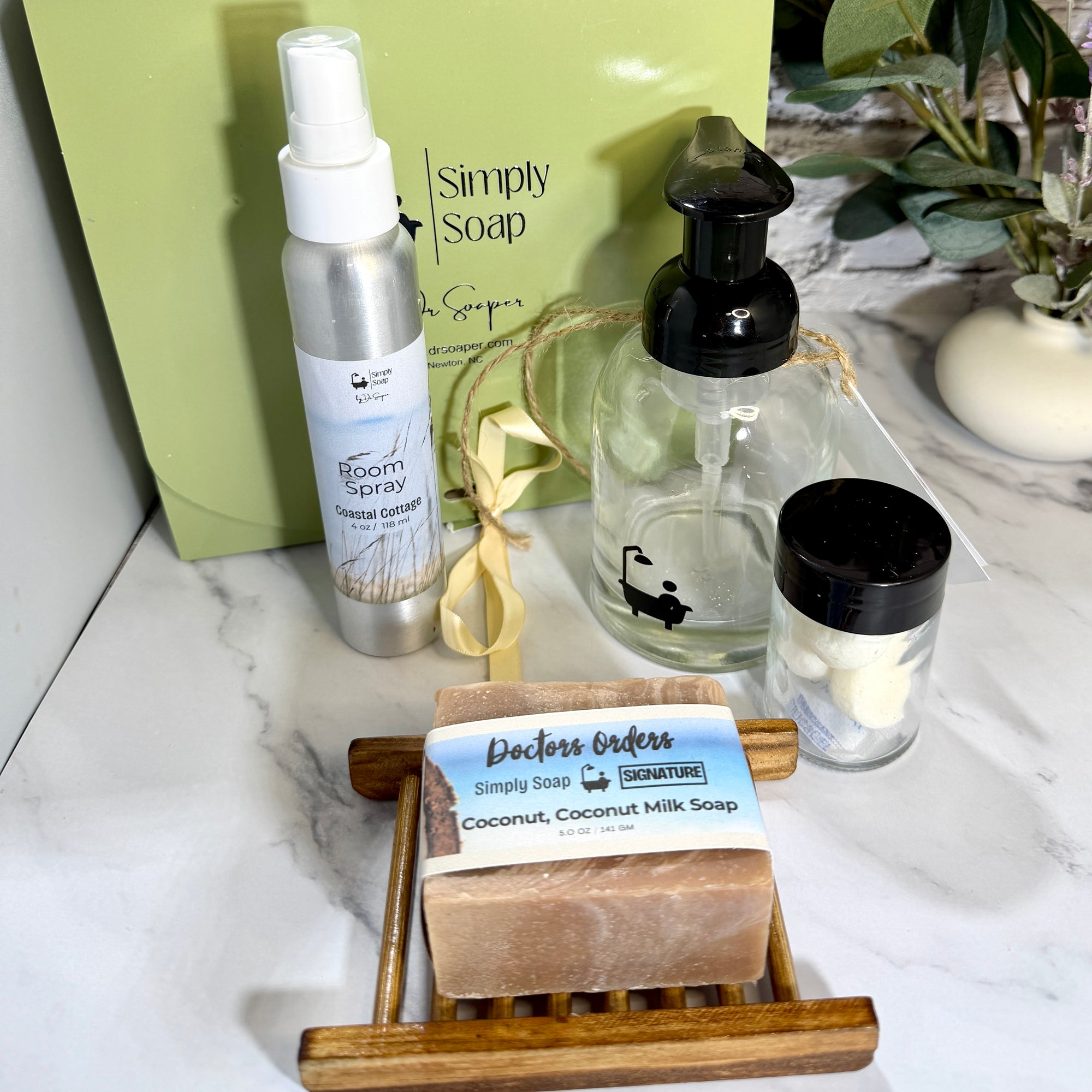 A curated bath and body gift set from Simply Soap on a white marble surface, featuring a silver room spray, a clear pump bottle, a small jar of soap tabs, and a bar of soap on a wooden dish,  with a green Simply Soap box in the background.