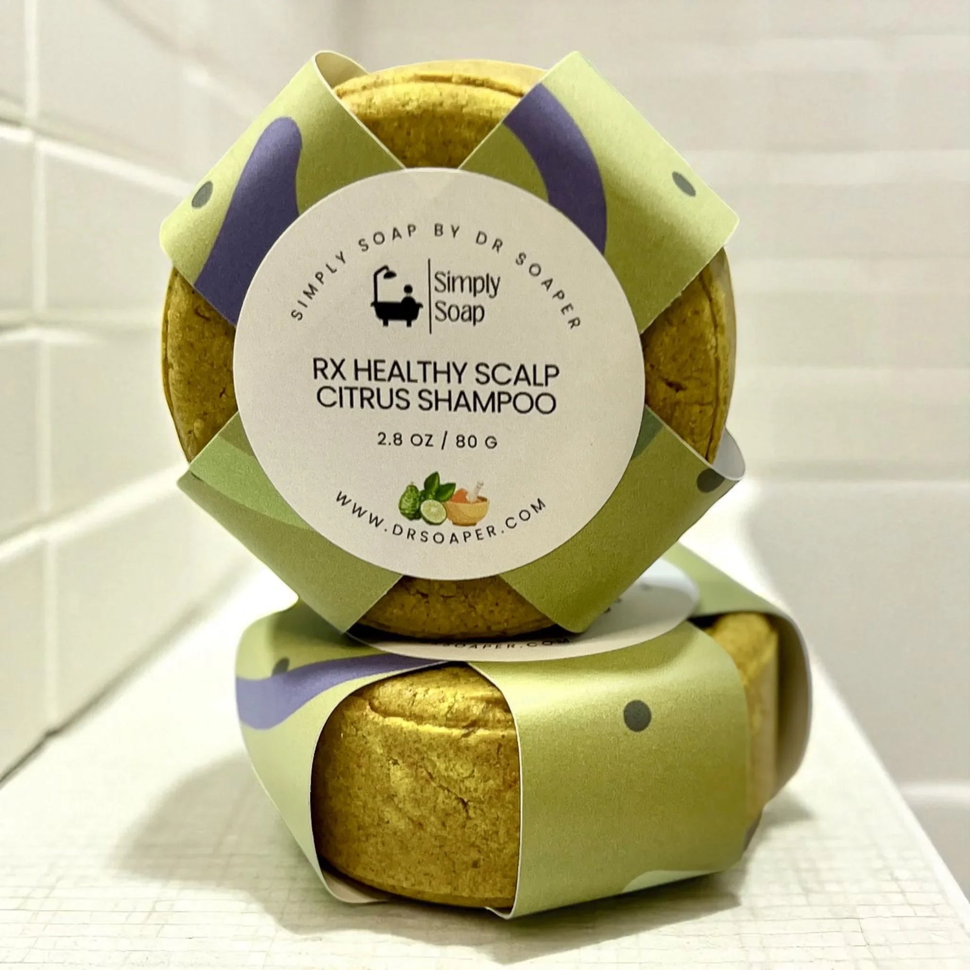Two bars of soap wrapped in green and purple paper with a label on a white surface.