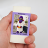 A hand holding a small cream-colored soap bar wrapped in a purple label with SUDZ in a robe