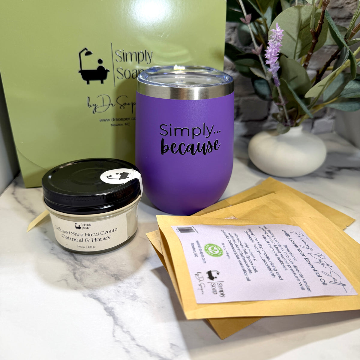 A gift set from Simply Soap by Dr. Soaper on a white marble surface, featuring a purple insulated tumbler with 'Simply because' printed on it, a jar of oatmeal and honey hand cream,  4 individual use bath salt sachets in a light green gift box. 