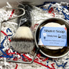 Shave brush and soap in a container on shredded paper with a visible brand label.