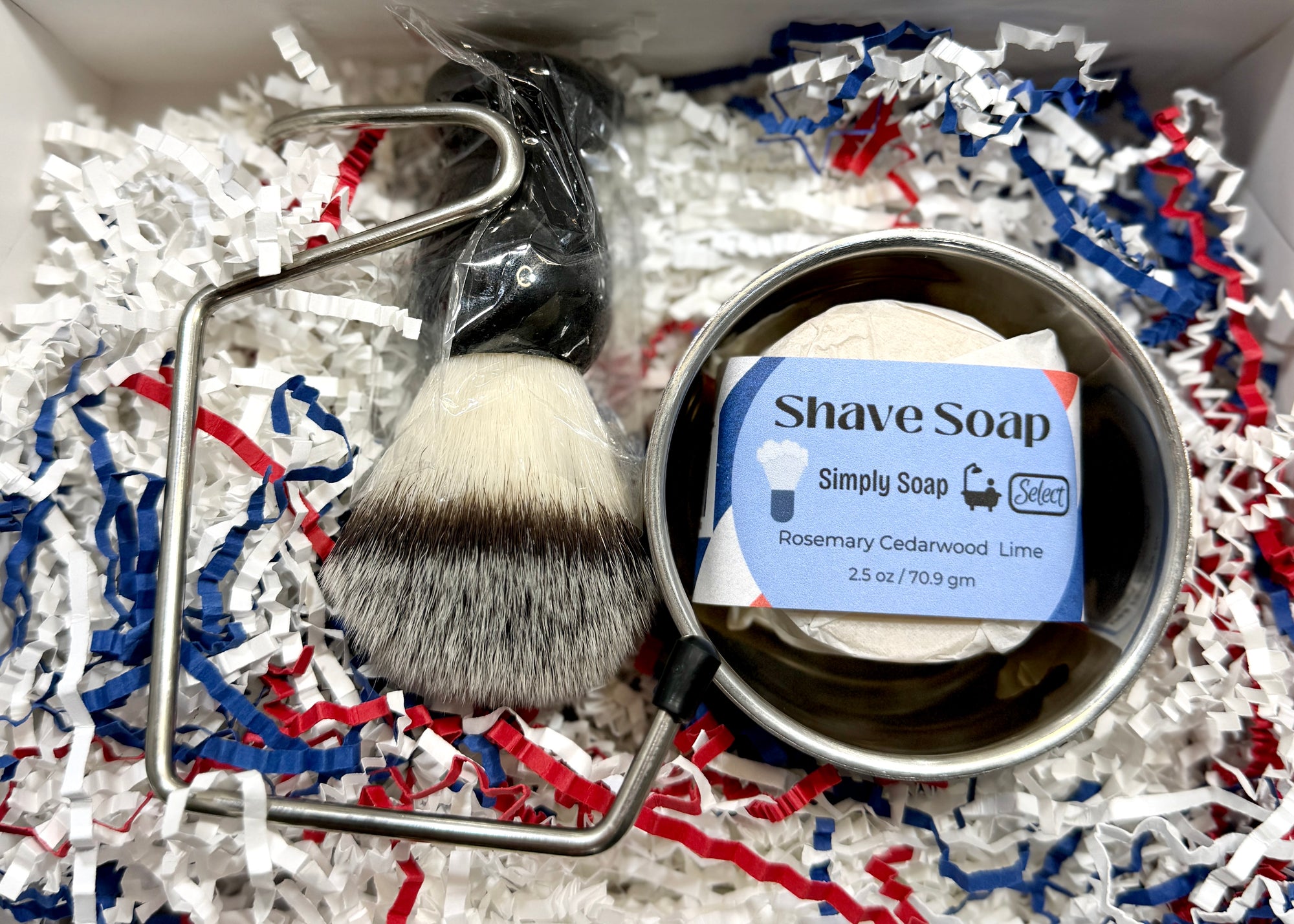 Shave brush and soap in a container on shredded paper with a visible brand label.