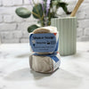 Two bars of shave soap in packaging on a marble surface with a plant in the background.