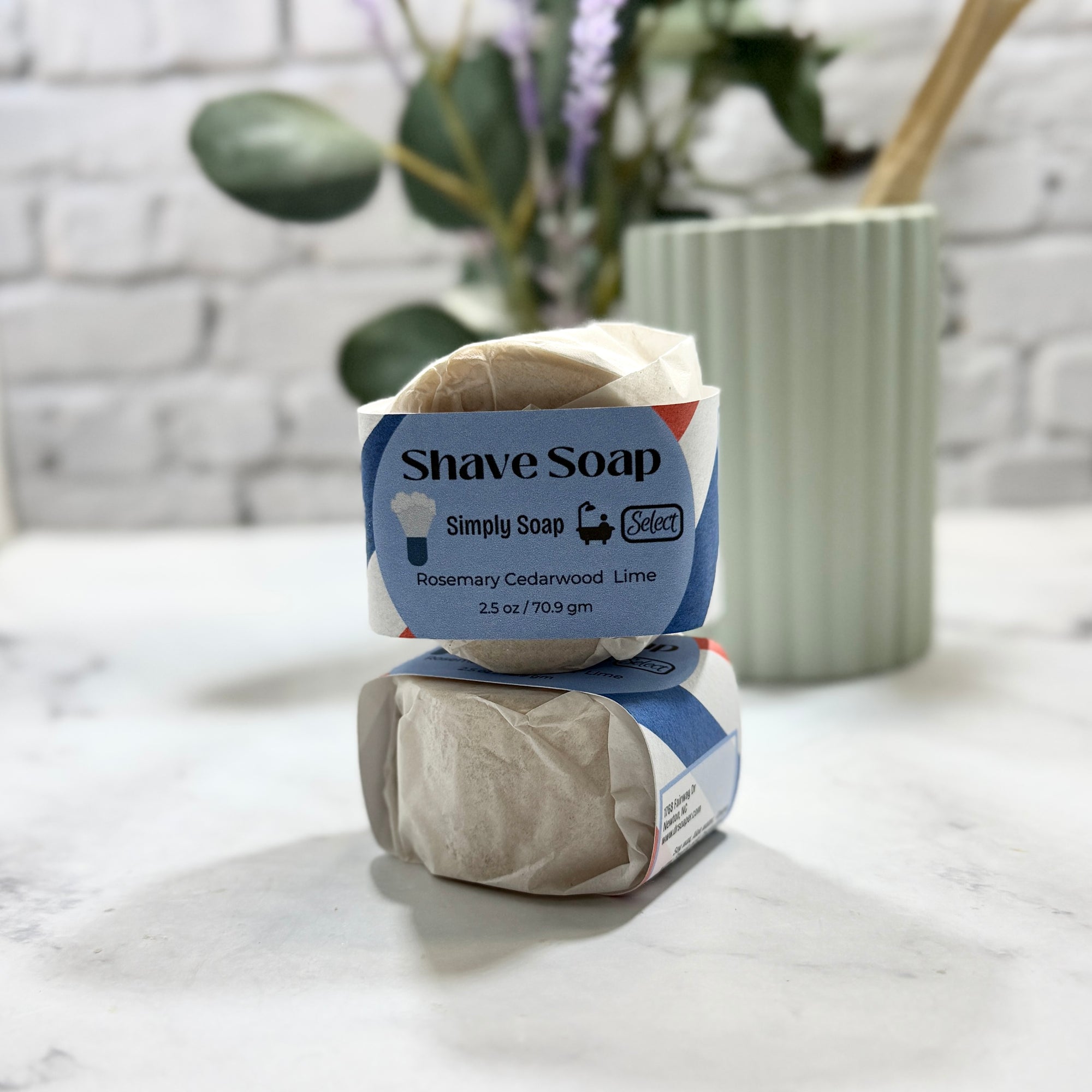 Two bars of shave soap in packaging on a marble surface with a plant in the background.