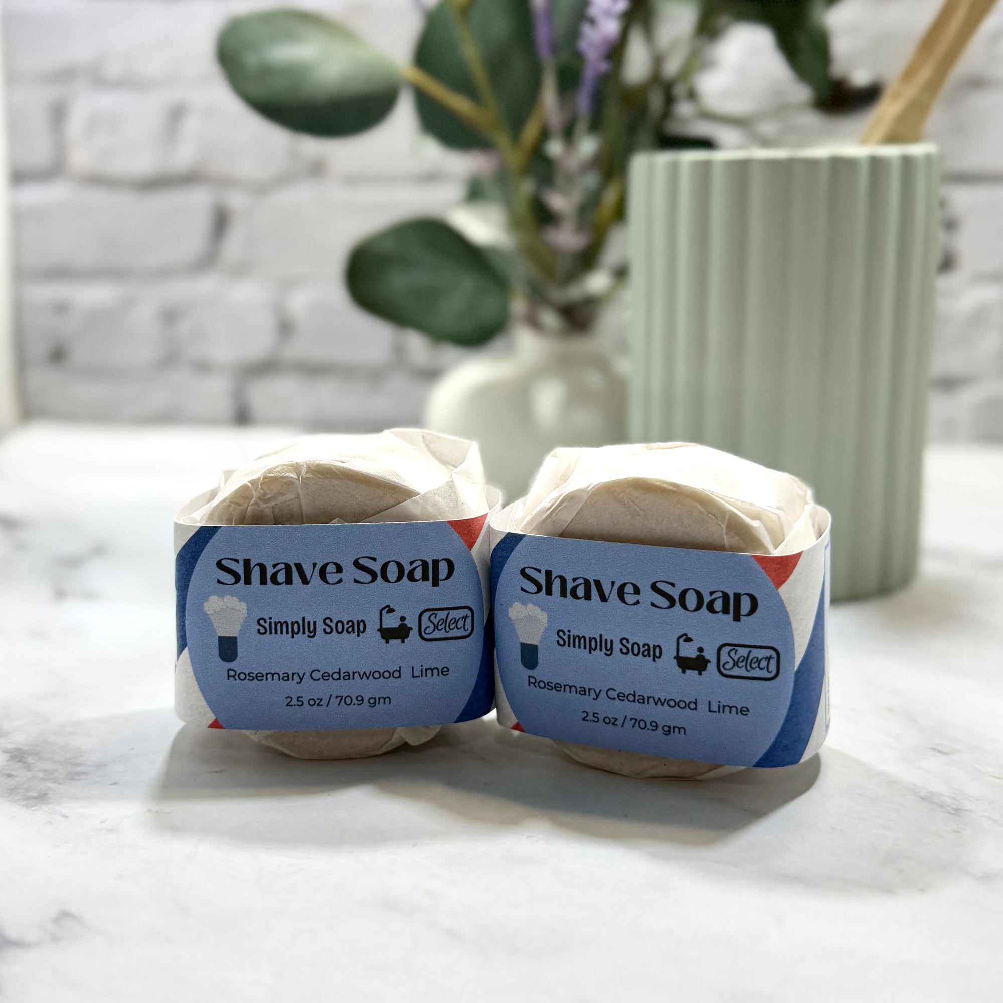 Two bars of shave soap in packaging on a marble surface with a plant in the background.