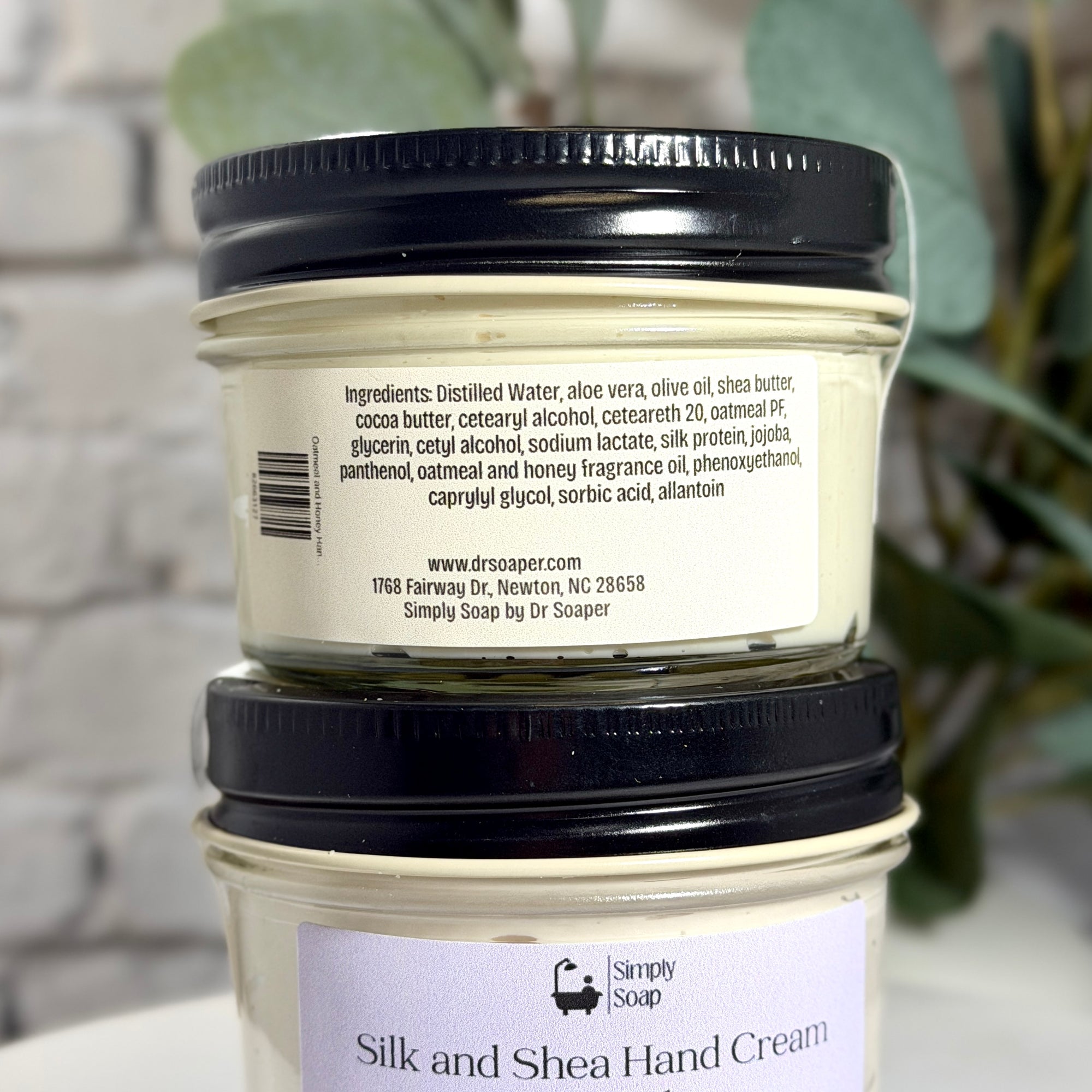 Two stacked jars of Simply Soap Silk and Shea Hand Cream, showing the back label of the top jar and the front label of the bottom jar against a soft-focus background.