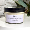 A jar of Simply Soap Silk and Shea Hand Cream in Lavender scent, featuring a black lid and a light lavender label, displayed on a white surface with a blurred brick wall and green leaves in the background.