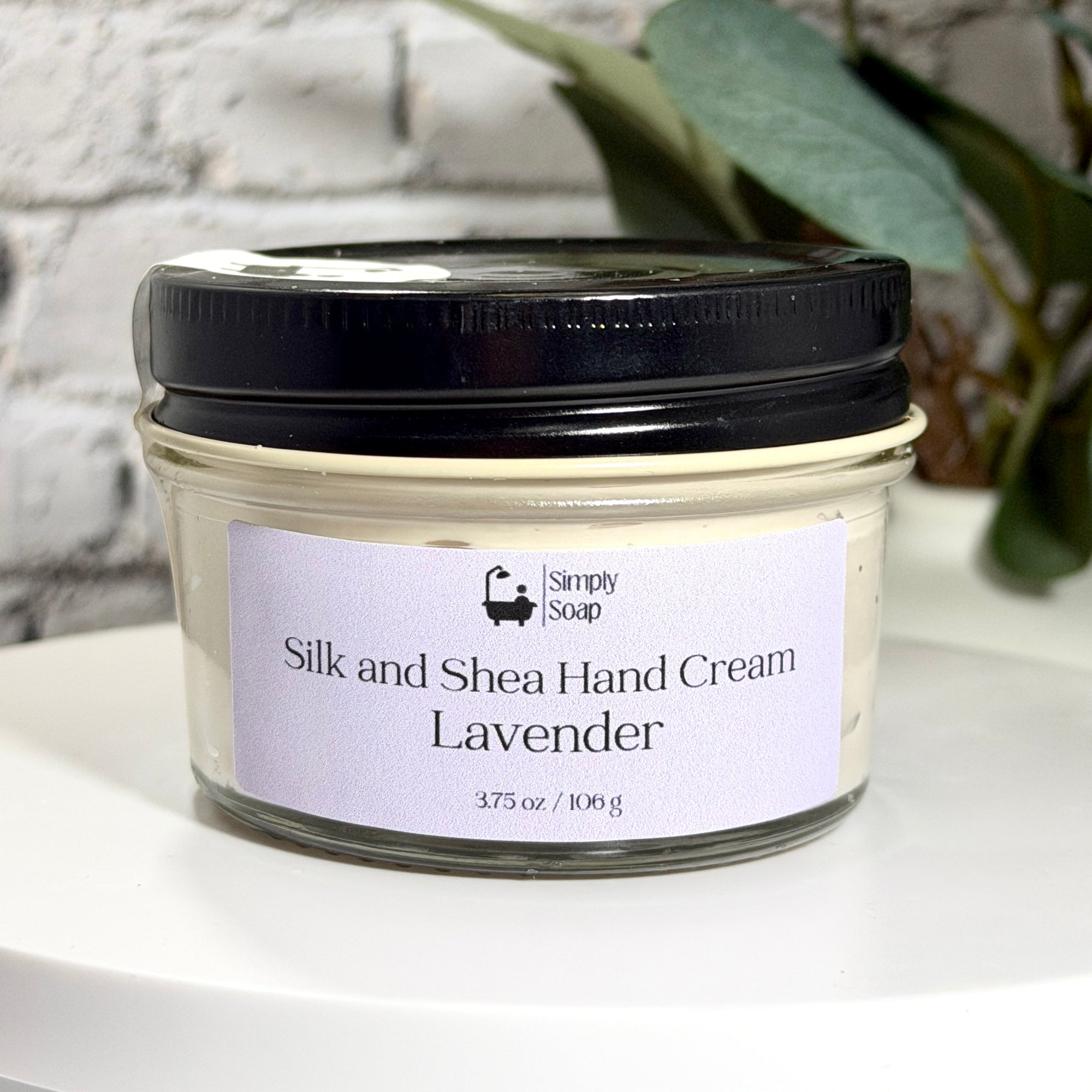 A jar of Simply Soap Silk and Shea Hand Cream in Lavender scent, featuring a black lid and a light lavender label, displayed on a white surface with a blurred brick wall and green leaves in the background.