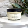 A glass jar of Simply Soap Silk and Shea Hand Cream with oatmeal and honey scent, featuring a cream-colored label and a black lid, displayed on a white pedestal with a blurred white brick wall and green leaves in the background.