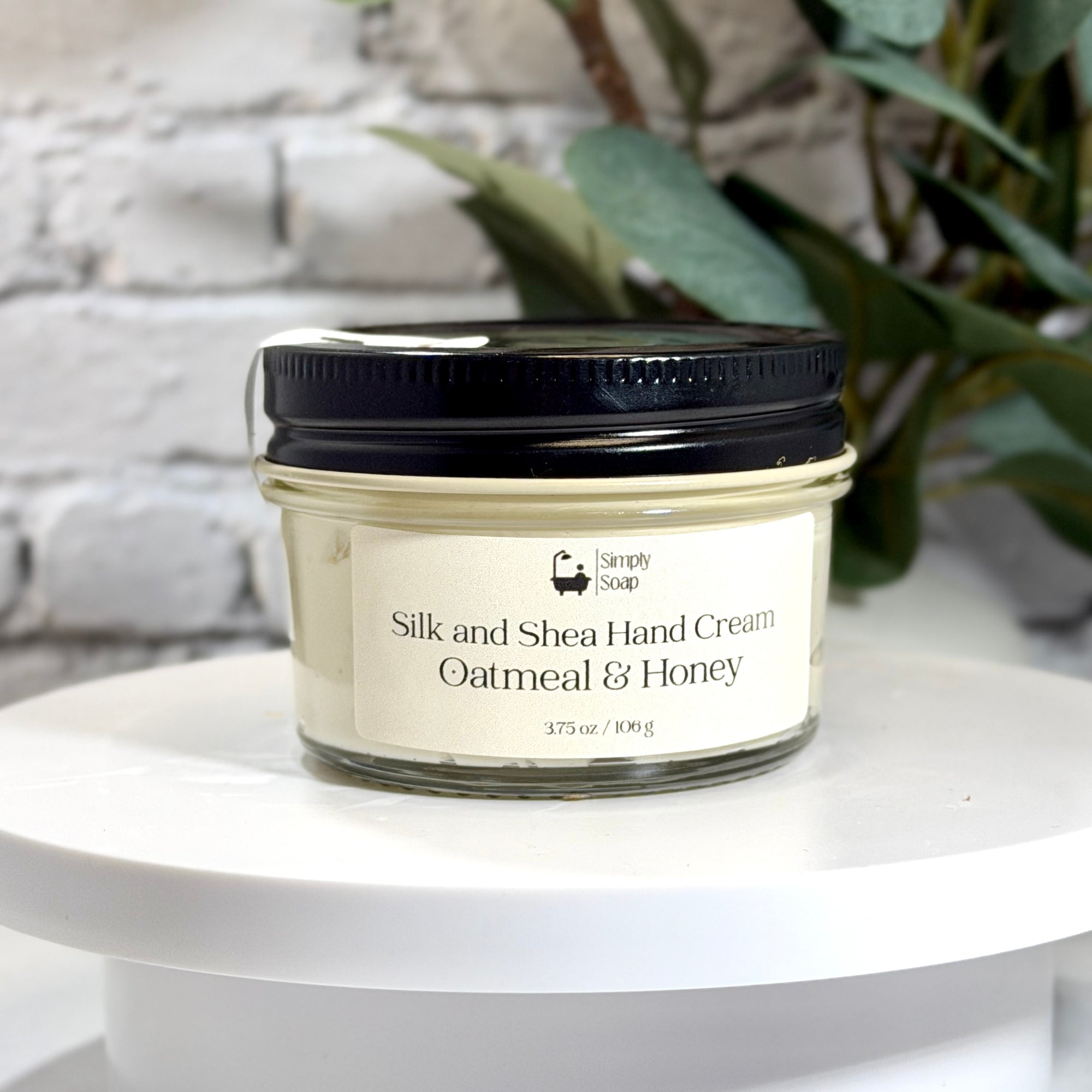 A glass jar of Simply Soap Silk and Shea Hand Cream with oatmeal and honey scent, featuring a cream-colored label and a black lid, displayed on a white pedestal with a blurred white brick wall and green leaves in the background.
