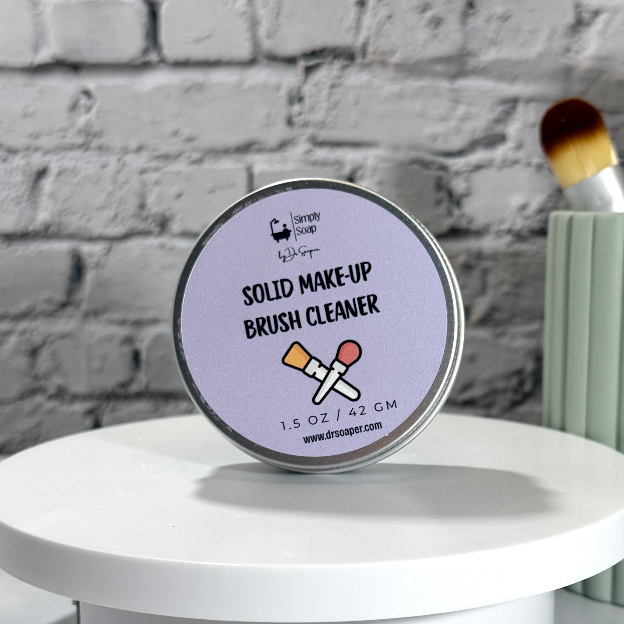 A round metal tin of Dr. Soaper's 'Solid Make-Up Brush Cleaner' displayed on a white round pedestal against a gray brick wall background.