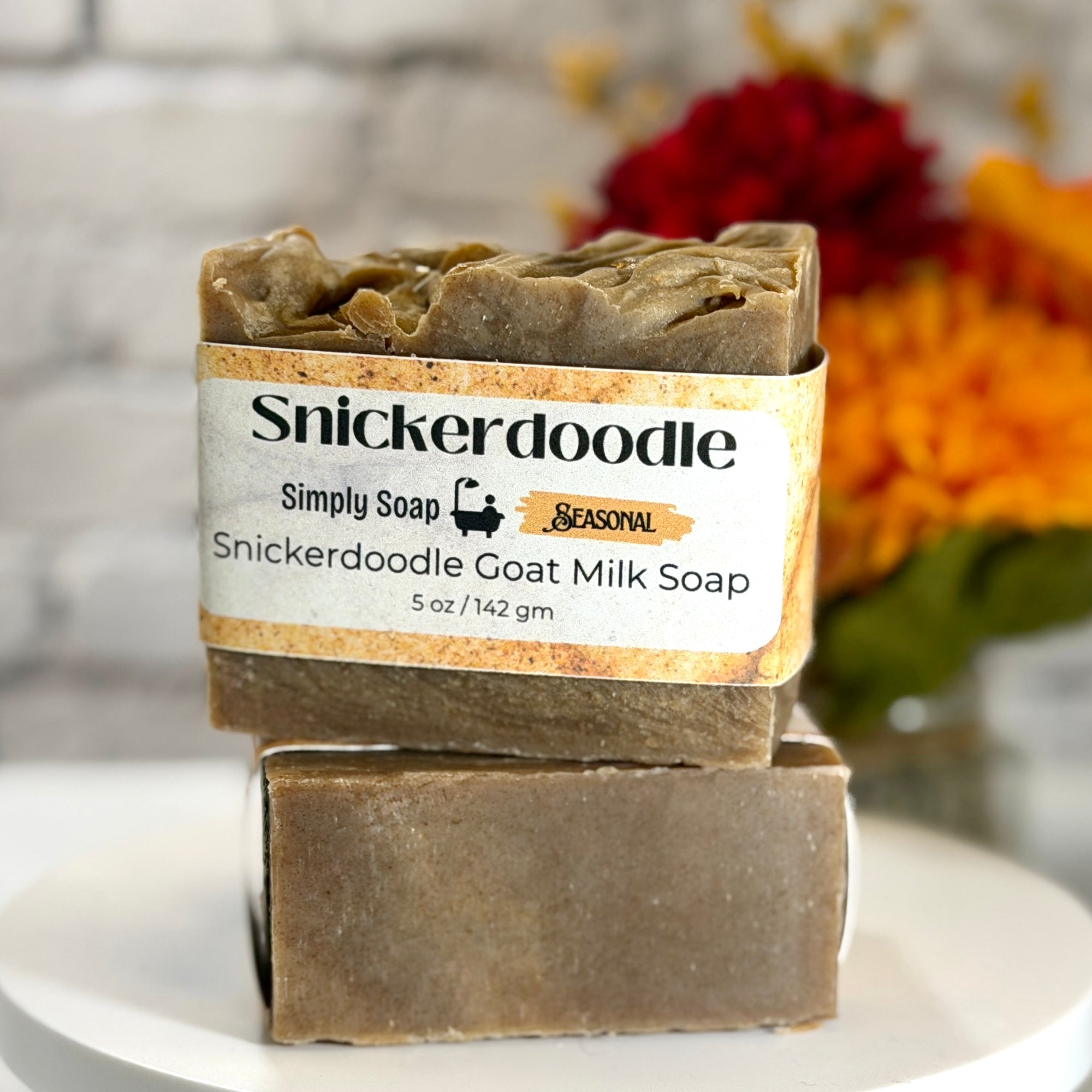 Two bars of Snickerdoodle Goat Milk Soap stacked on a white plate, with a paper label wrapped around the top bar. The background shows a blurred white brick wall and a vase of colorful flowers.
