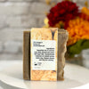 A handmade soap bar wrapped in a kraft paper band with a white product label, displayed on a white surface with a blurred background of flowers.