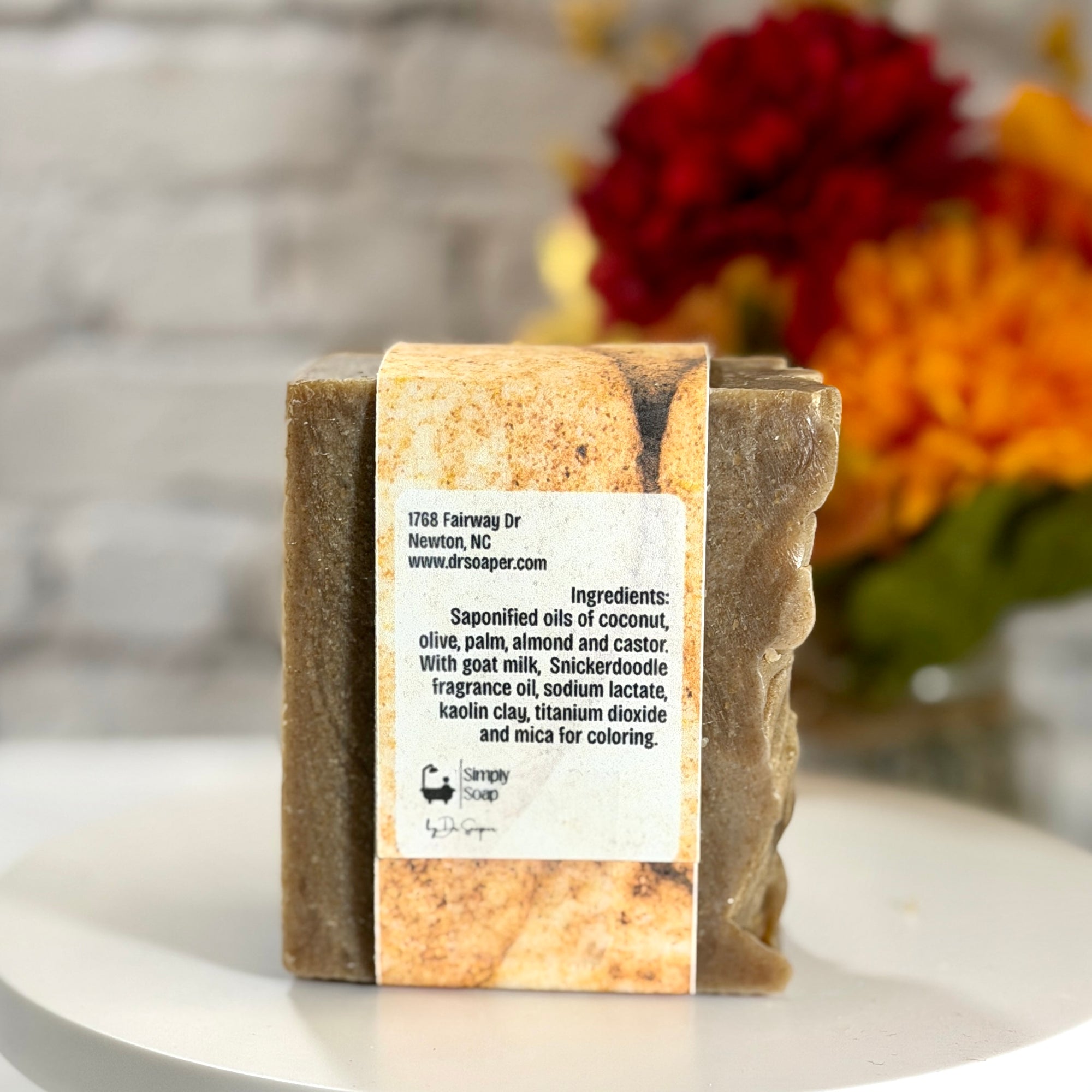 A handmade soap bar wrapped in a kraft paper band with a white product label, displayed on a white surface with a blurred background of flowers.