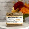 A bar of Snickerdoodle Goat Milk Soap by Simply Soap, wrapped in a white paper label with a rustic orange border, displayed on a white surface with a blurred background of flowers and a white brick wall.