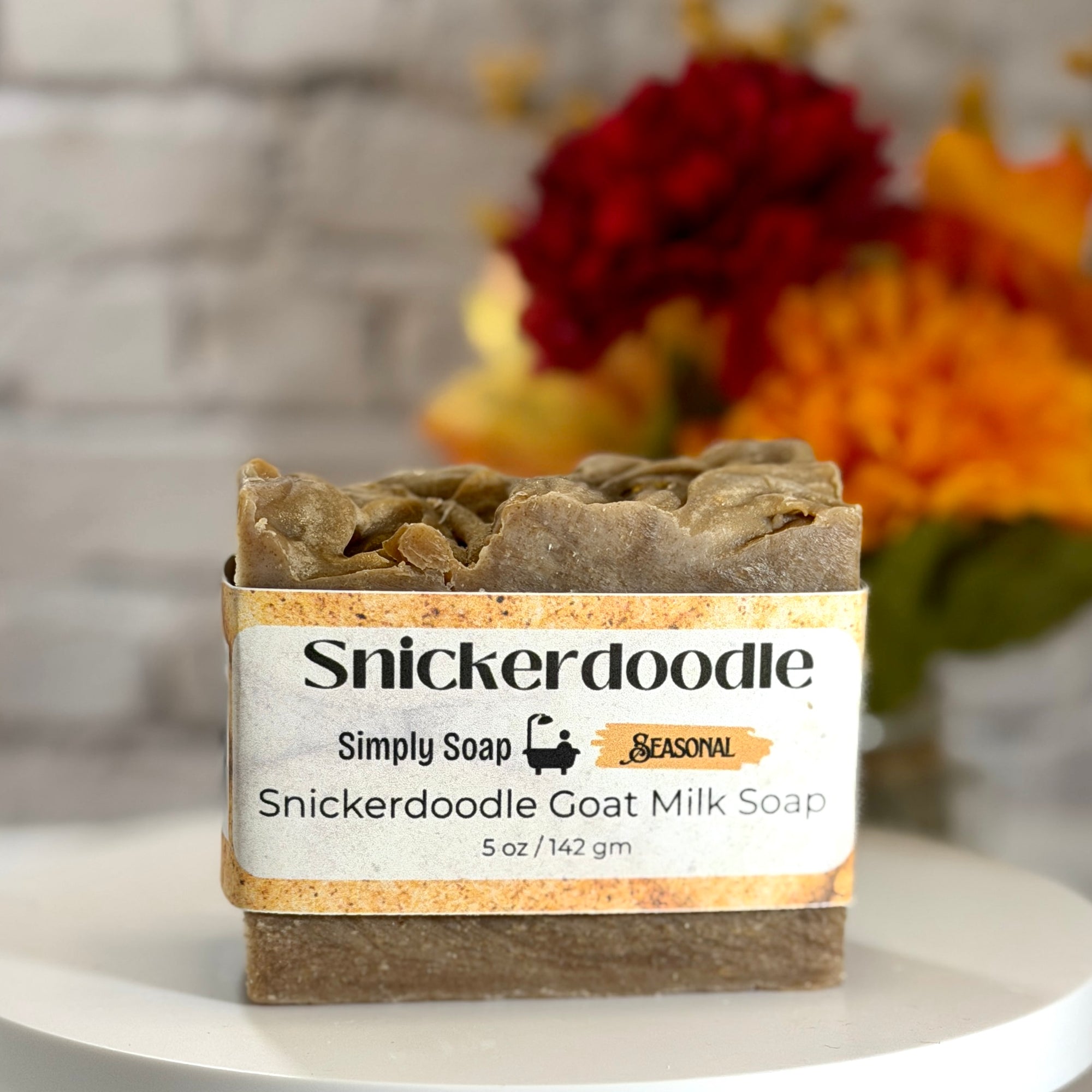 A bar of Snickerdoodle Goat Milk Soap by Simply Soap, wrapped in a white paper label with a rustic orange border, displayed on a white surface with a blurred background of flowers and a white brick wall.