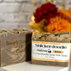 Two bars of Simply Soap Snickerdoodle Goat Milk Soap displayed on a white surface with a blurred brick wall and autumn flowers in the background.