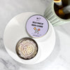 A round metal tin of Dr Soaper's  Solid Make-Up Brush Cleaner sits on a white plate on a marble surface. The lid is off, showing the pale lavender label with the product name, and the open tin reveals a decorative, flower-shaped solid makeup cleaner soap bar. 