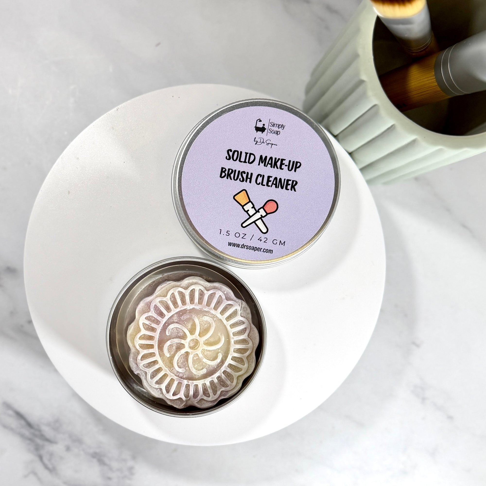 A round metal tin of Dr Soaper's  Solid Make-Up Brush Cleaner sits on a white plate on a marble surface. The lid is off, showing the pale lavender label with the product name, and the open tin reveals a decorative, flower-shaped solid makeup cleaner soap bar. 