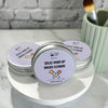 Three round metal tins of  Solid Make-Up Brush Cleaner arranged on a white round tray, with a mint green brush holder containing brushes in the background against a light brick wall.