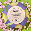 Simply Soap Bloom Essence Conditioner packaging surrounded by pink flowers on a green background