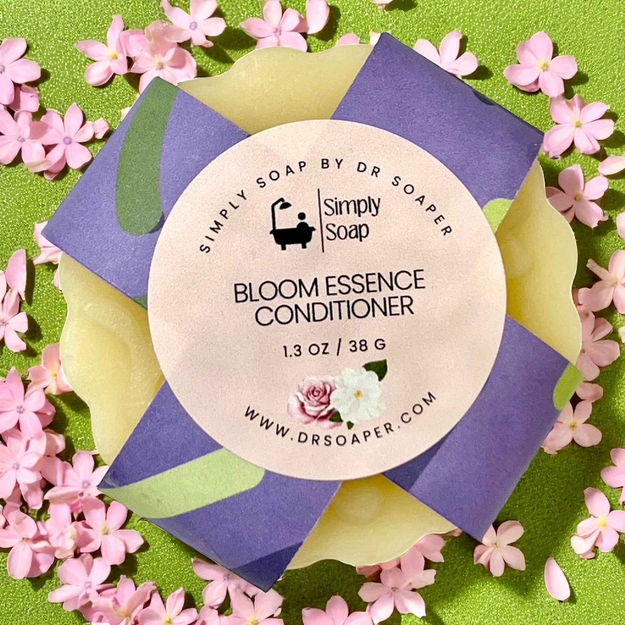 Simply Soap Bloom Essence Conditioner packaging surrounded by pink flowers on a green background