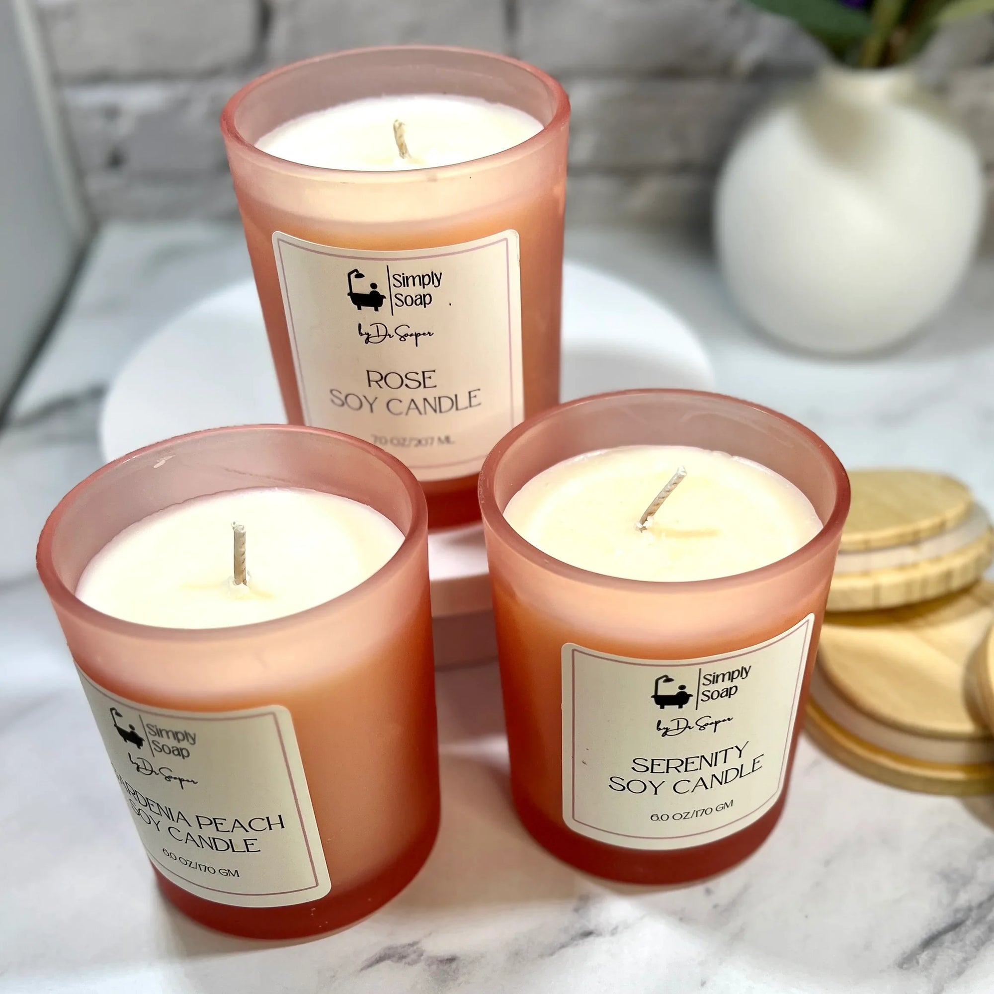 Three open soy candles by Dr Soaper, with wooden lids on a marble surface with a white brick wall background.