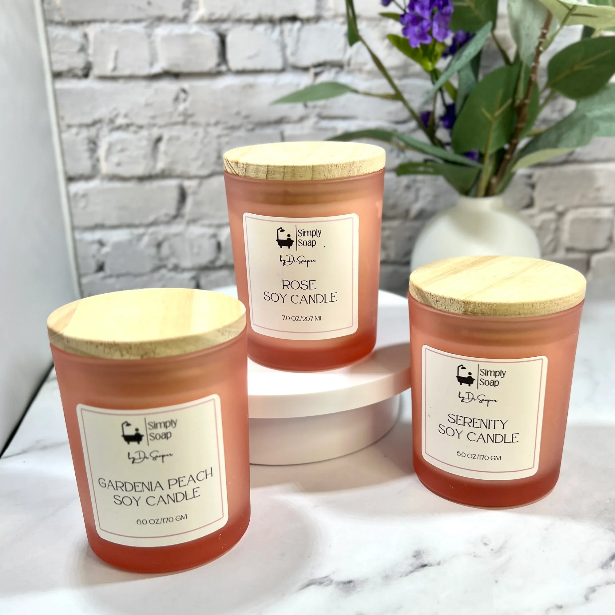 Three soy candles by Dr Soaper, with wooden lids on a marble surface with a white brick wall background.