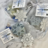 Snowflake-shaped soap bars in clear bags with decorative snowflake decorations on a marble surface.