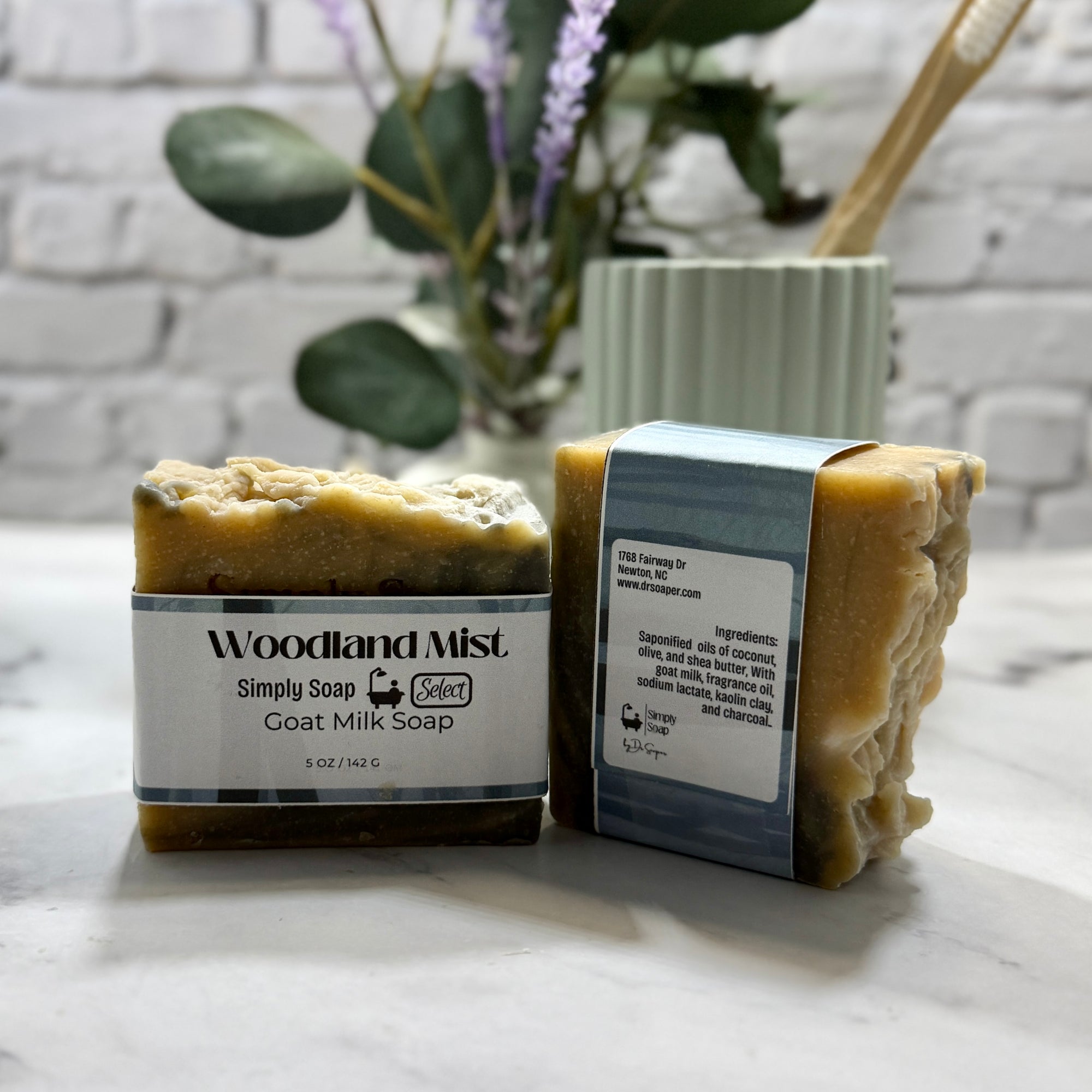 Two bars of Woodland Mist goat milk soap on a marble surface with a blurred plant in the background.