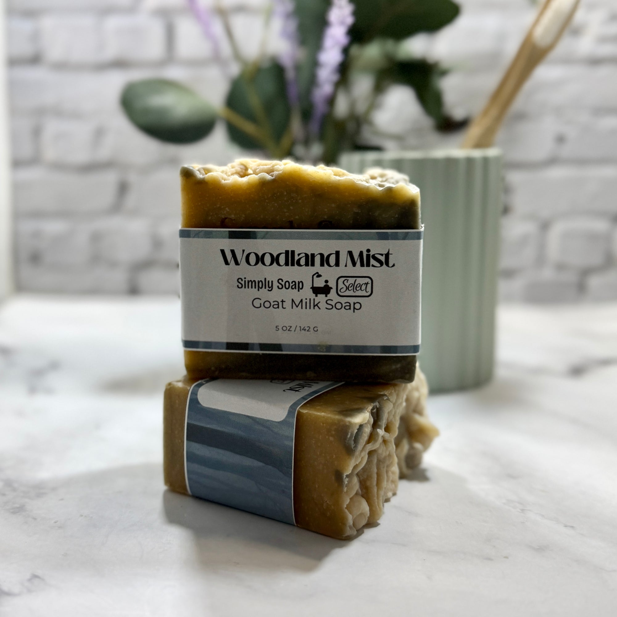 Two bars of Woodland Mist goat milk soap on a marble surface with a blurred plant and container in the background.